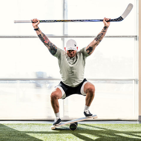 hockey player in gym with green turf floor squatting on balance board and holding hockey stick