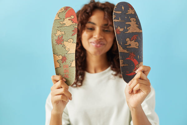 Woman with curly hair holding up 2 cork insoles. One is green and one is blue. The background is light blue. The girl is wearing a white shirt