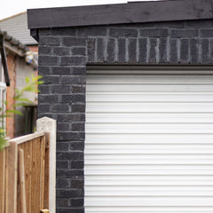 A garage with a white door, clad in black brick slips, with a wooden fence and facia.