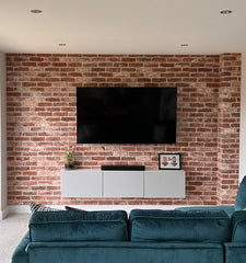 A bright reclaimed red brick feature wall with a central TV, and a wall unit underneath in grey. the top of a blue velvet sofa is in shot at the bottom.