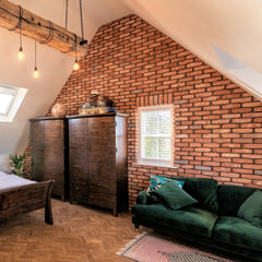 A gable end is clad is light red brick slips with blue accents. There are 2 mahogany wardrobes to the left of a small central window, and a green velvet sofa to the right, all in front of a parquet floor.