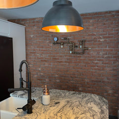 A red brick feature wall in a kitchen, a marbled worktop is at the bottom of the picture with a black top showing. There is a bowl light shade hanging from the ceiling, and on the brick slip wall is copper piping with lightbulbs and a pressure gauge at the ends.