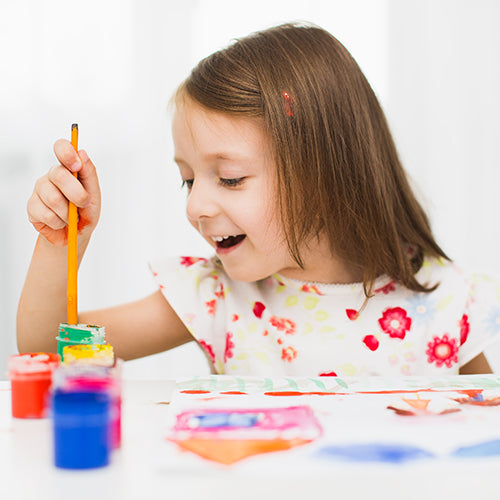 Young girl sitting at white table painting with multiple paint colors and brush