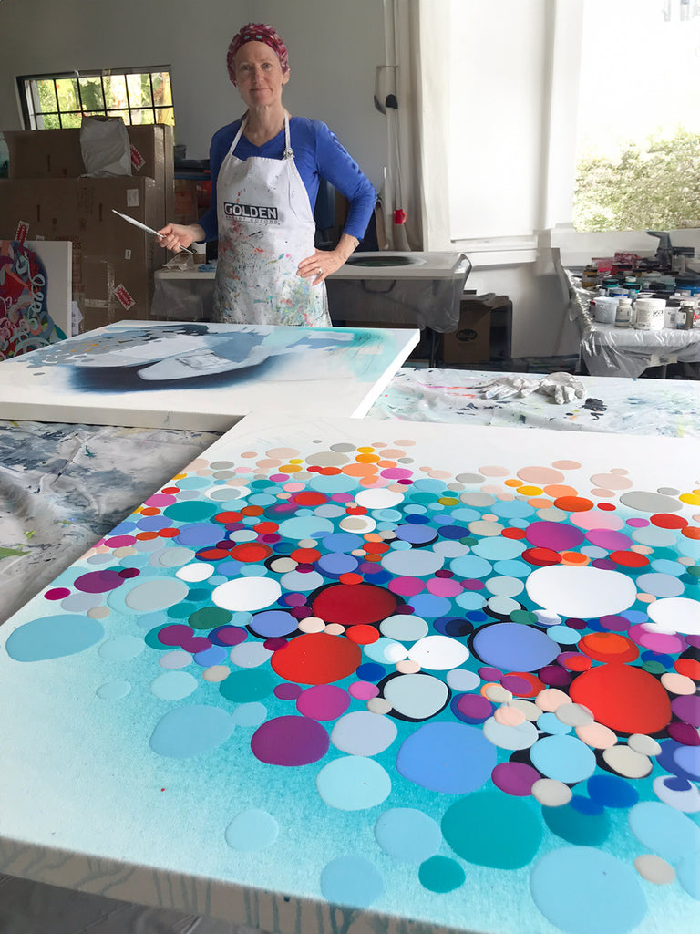 Artist Claire Desjardins, stands before her painting table, paintbrush in hand.