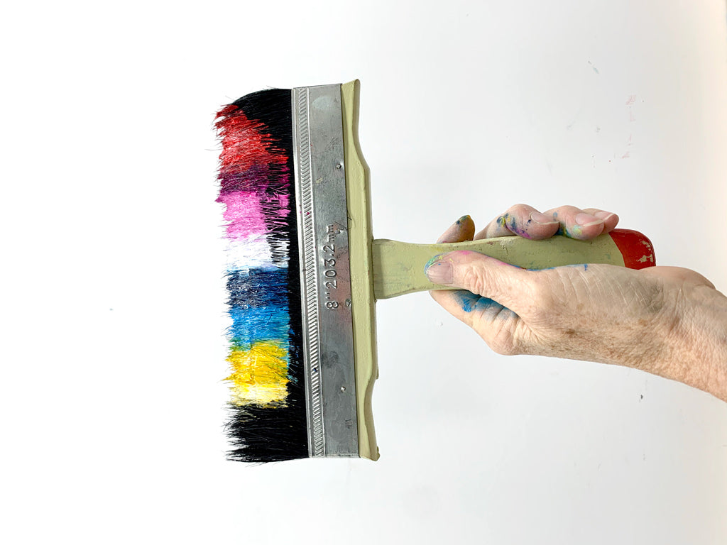 Wide artist paintbrush loaded with pink, blue, white and yellow acrylic paints.