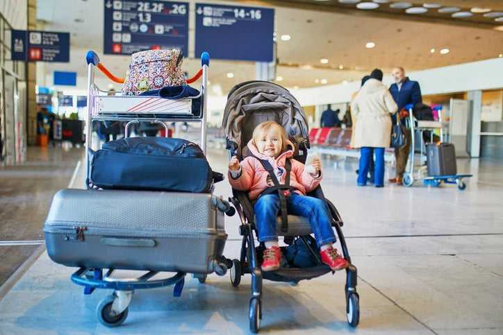 Baby girl in a travel stroller next to luggage and a luggage cart in a busy airport
