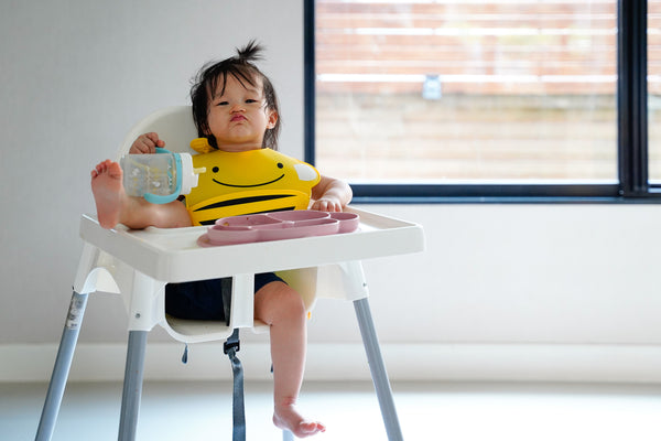 Baby girl eating on the best highchair and holding a water bottle by her hand while wearing a yellow bib, with her foot resting on the highchair tray