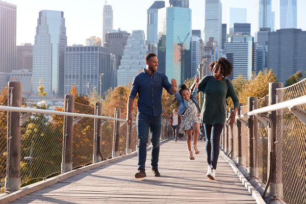 NYC mom and NYC dad and young daughter holding hands walking across a footbridge with a major city skyline in the background