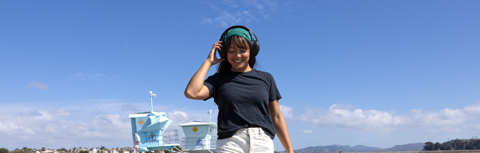 : A smiling women in a black t-shirt, strolling on the beach adjusts the volume using the on-ear push-button control on her REVO wireless TW340 headphones from TWT Audio.
