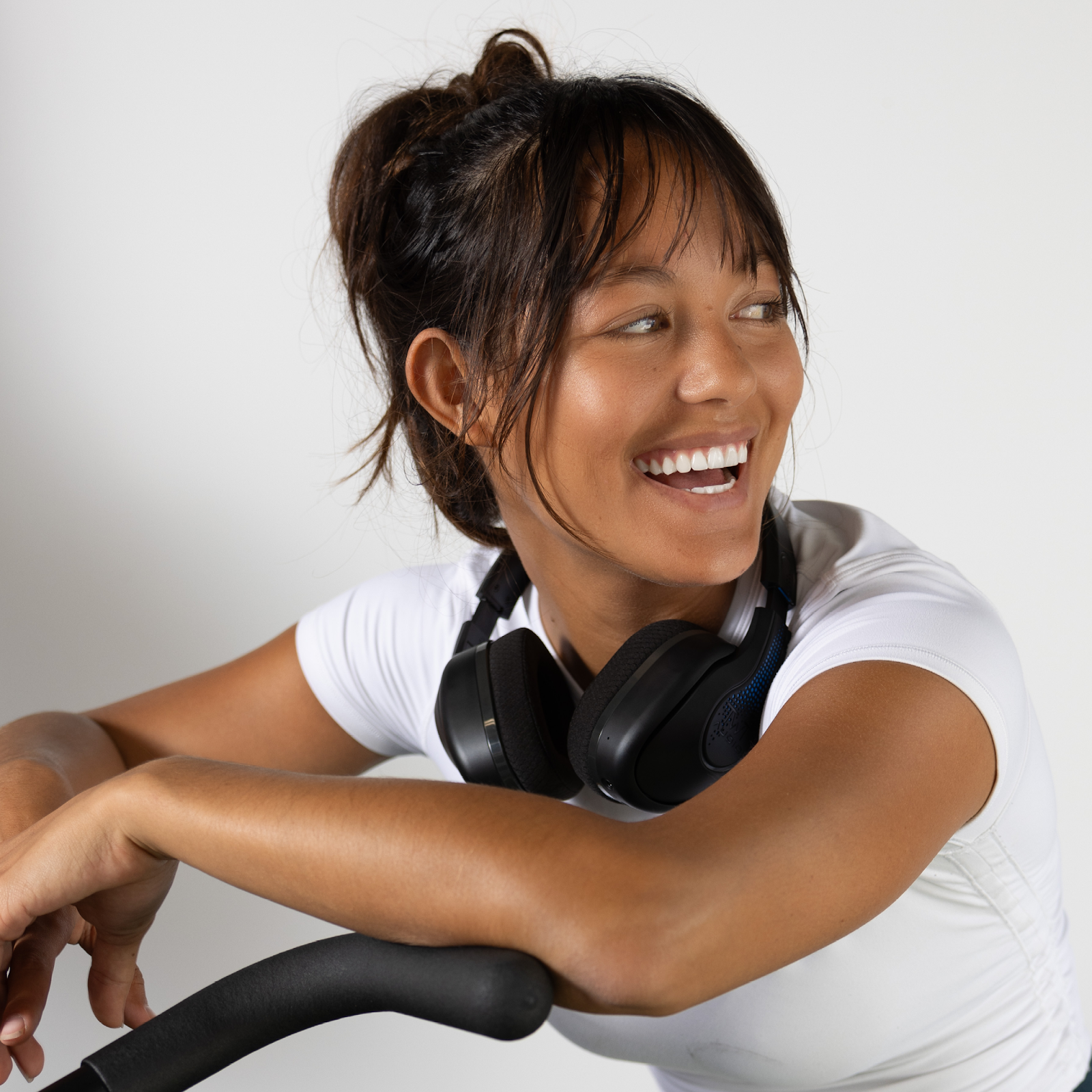 A woman, with a smile directed towards someone on her left, casually rests her TW340 headphones from TWT Audio around her neck while seated on a stationary bike, enjoying her workout.
