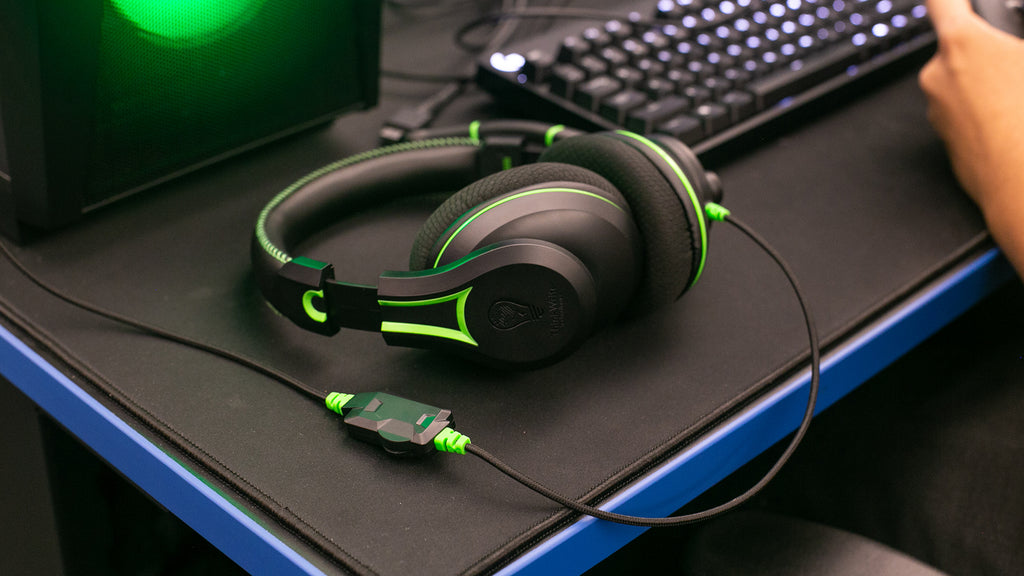 The 250XG Victory Gaming Headset by TWT Audio resting on a desk in a classroom, positioned in front of a gaming PC. The headset is showcased with a focus on its volume and inline gaming-style volume control with mic mute functionality.