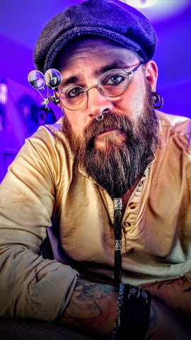 Man with long braided beard wearing glassess with attached small magnifying lenses, tan tunic and flat cap. Leaning by his elbows onto a desk.