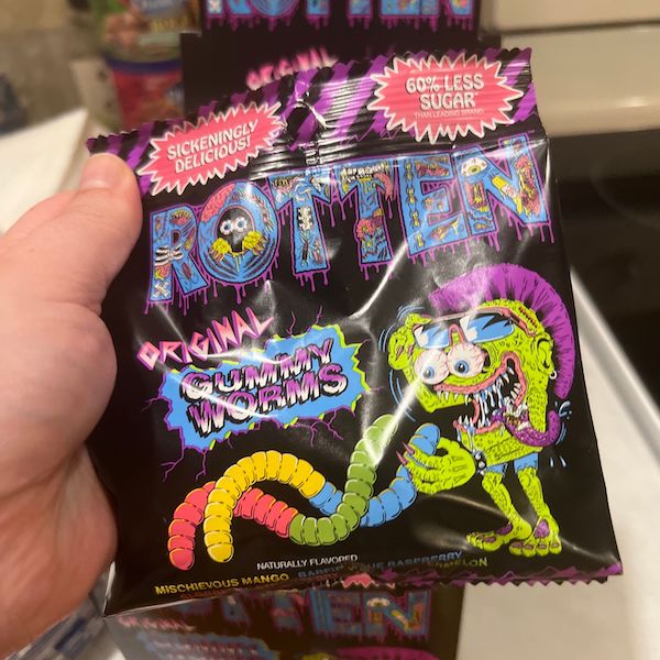 A hand holding a colorful packet of 'Rotten Gummy Worms' candy with funky cartoon graphics.