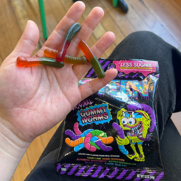 Hand holding gummy worms over an open candy bag labeled 'Gummy Worms'.