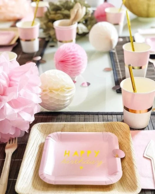 Tischdekoration für einen Geburtstag in Rosa mit 'Happy Birthday' Pappteller auf Palmblatter, Holzbesteck, rosa und weißen Wabenbälle, rosa Pom Pom und rosa, weiß, goldenen Pappbechern und goldenen Papierstrohhalmen