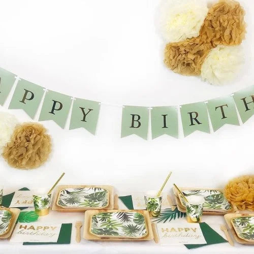Partydekoration mit grüner 'Happy Birthday' Girlande, weißen und goldenen Pom Poms, Palmblatt geschirr, 'HAPPY birthday' Servietten und Holzbesteck