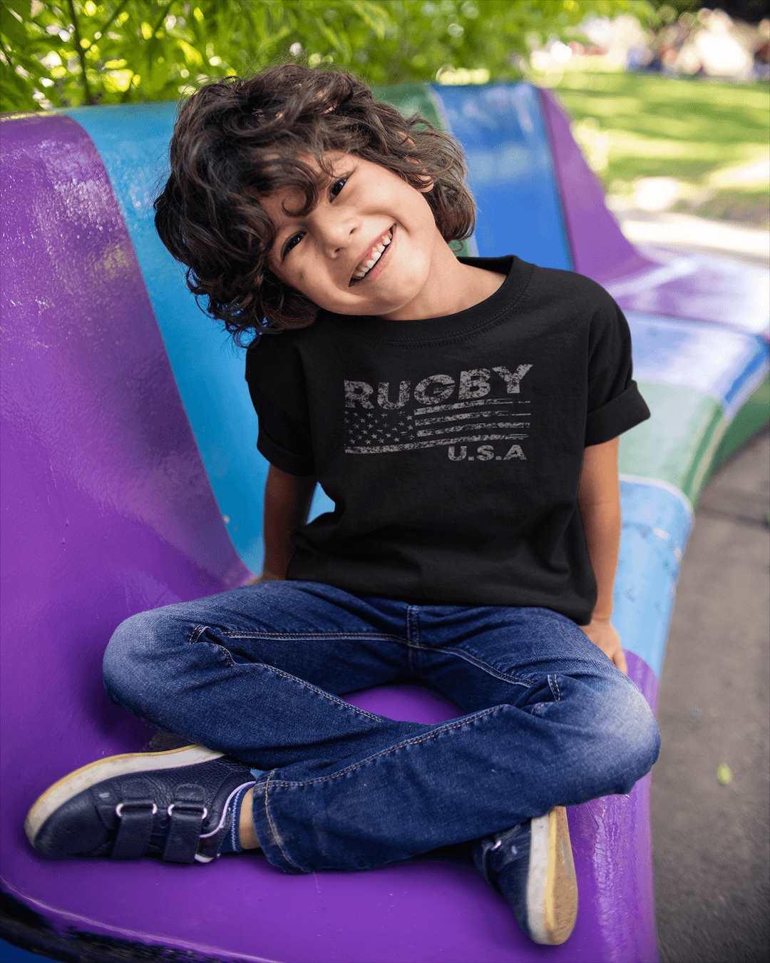 RUGBYNOMAD black organic cotton kids classic streetwear t shirt with south africa springbok artistic design, boy model, sitting on purple blue bench, smiling, black jeans, tennis shoes Edit