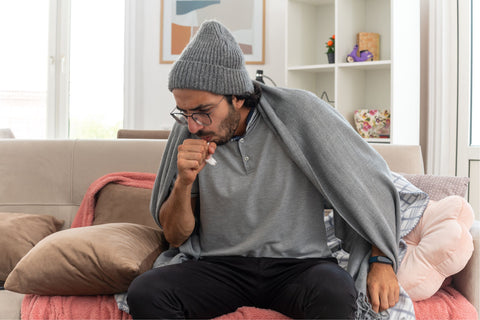 Sick young caucasian man in optical glasses wearing winter hat coughing keeping fist close to mouth sitting on couch at living room