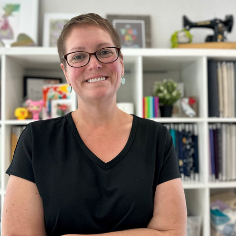 Michelle Adcock, owner of Miche Niche. Wearing a black dress with arms crossed in front of a bookshelf of colorful fabric lined on shelves.