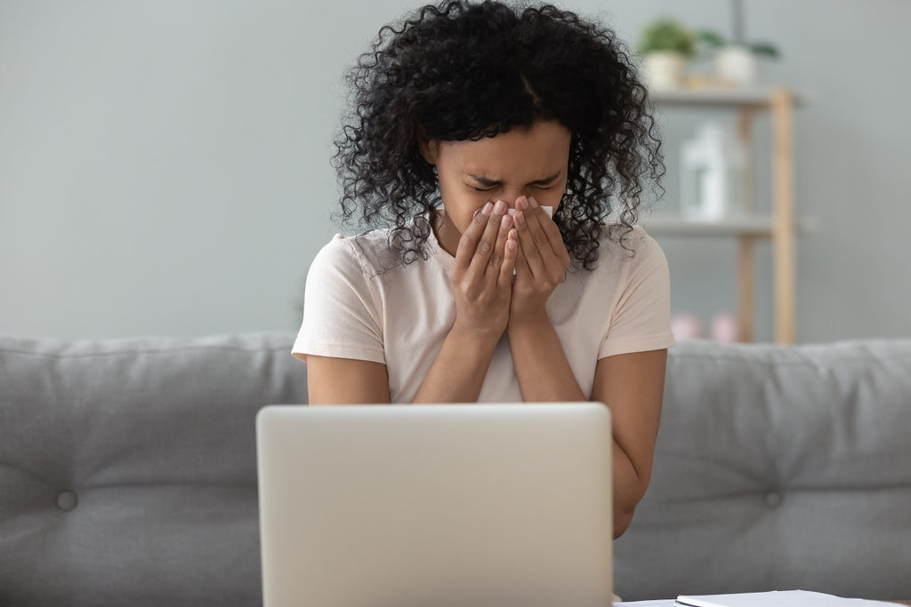 Person sitting on couch in front of laptop blowing nose due to allergies.