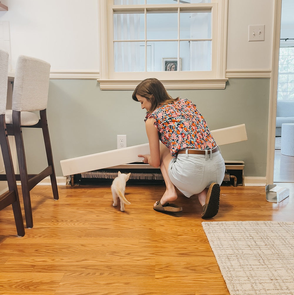 Person removing the old cover on a baseboard heater with a kitten in the foreground.