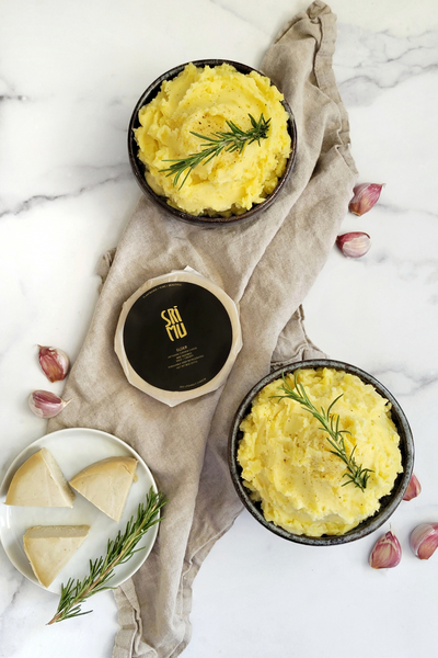 Two bowls of mashed potatoes seen from above, with a cheese wheel and ingredients spread out around it