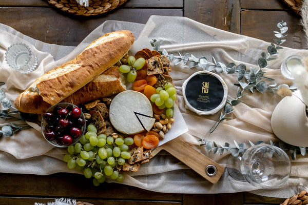 table decorated with cheese board bread fruits and cheese wheels seen from above 