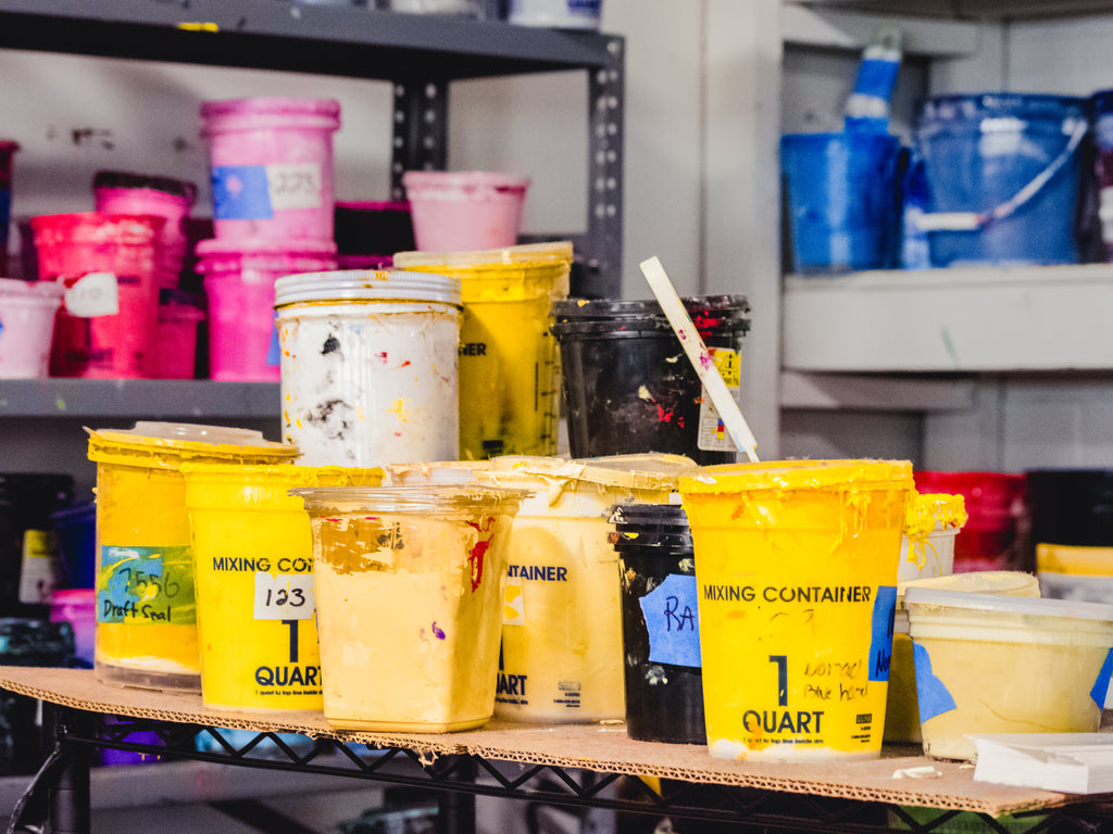 Buckets of yellow screen printing inks, center front and in focus. In background right are buckets of blue screen printing ink. In background left are different shades of pink screen printing ink.