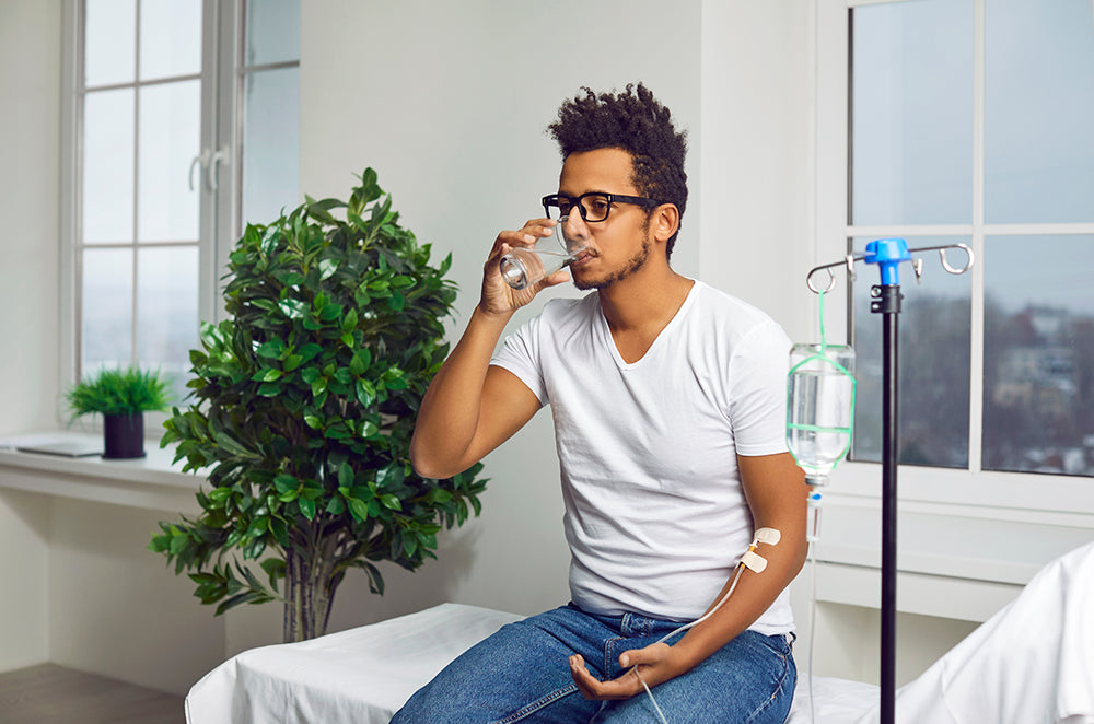 Young man drinks a glass of water while receiving mobile IV therapy.