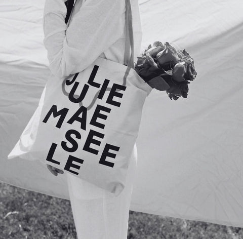 A model holding a tote bag that reads "Julie Maeseele" with flowers coming out of the bag