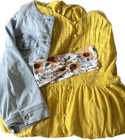 A yellow sundress with a light blue jean jacket over it and a bright and playful sunflower print headband laid across it. 