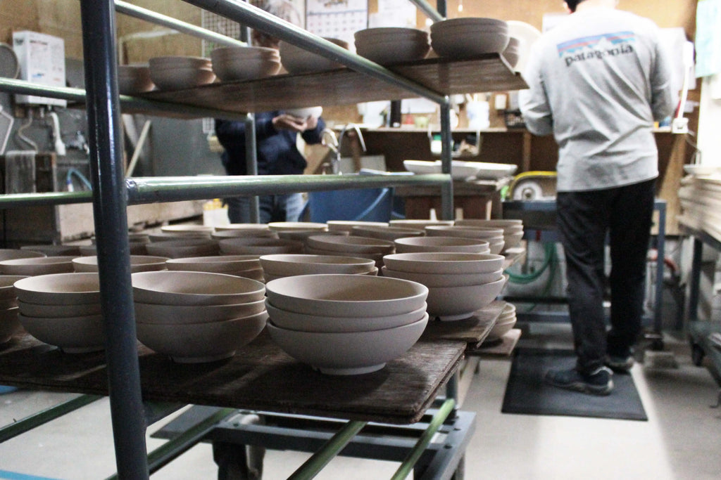 工房内で積み重ねられた未焼きの陶器のボウルと、背景に作業をする職人の様子