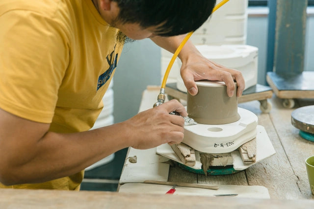 かたむけてねこ茶碗を石膏型からコンプレッサーを使って取り出している工房の風景。作業者は黄色のTシャツを着て作業に取り組んでおり、工房の作業台には陶器作りに使われる様々な道具が置かれている。