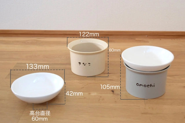 木の床に置かれたねこ茶碗３つの寸法を示す画像。左側の白い平らなねこ茶碗の直径は133mm、高さは60mm。中央のベージュ色のねこ台座の直径は122mm、高さは80mmで、「きなこ」とラベルが付いている。右側の灰色のねこ茶碗台座セットの直径は105mm、高さは42mmで、台座に「omochi」とラベルが付いている。各寸法は白い文字と数字で注記されている。
