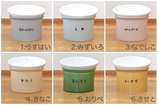 六つの異なる色の名入れされた台座とねこ茶碗が木製のテーブルの上に並べられた画像。左上から「omochi」と名入れされたグレーの台座は「1.うすはい」という説明が、中央上の「レオ」と名入れされた水色の台座には「2.みずいろ」と、右上の「momo」と名入れされたピンクの台座には「3.なでしこ」と書かれている。左下の「きなこ」と名入れされたベージュの台座には「4.きなこ」、中央下の「おりべ」と名入れされた緑の台座には「5.おりべ」、右下の「いくら」と名入れされたイエローの台座には「6.きせと」という説明がそれぞれ付いている。