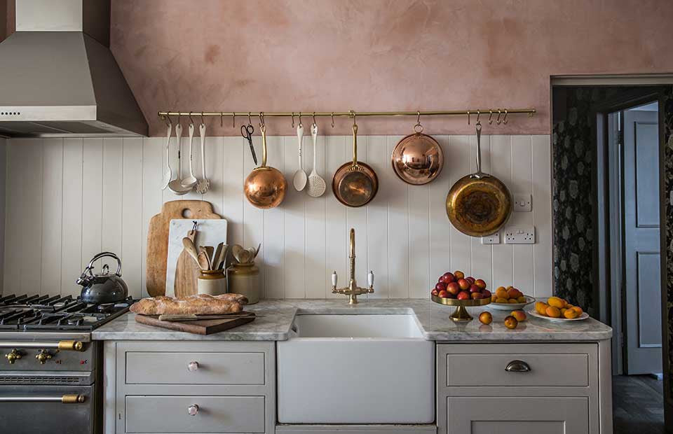 Cuisine avec un évier style farmhouse, cabinets crème, mur en tadelact rose, et une barre en laiton pour tenir les cuillères. 