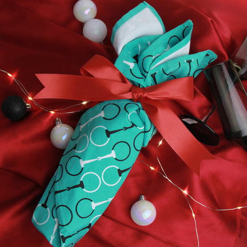 A bottle of wine wrapped in an equestrian tea towel with a red ribbon on a festive red satin background with lights and chrstmas ornaments