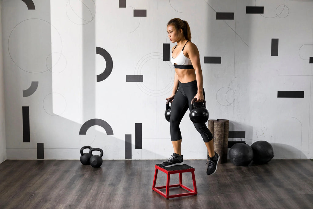 Fit Asian woman in workout clothes doing step-ups while holding a kettlebell in each hand