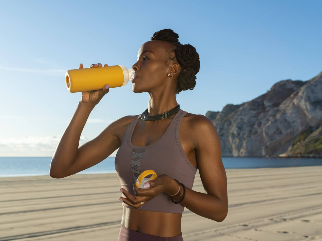 Fit black woman in a workout top drinking from a waterbottle on the beach