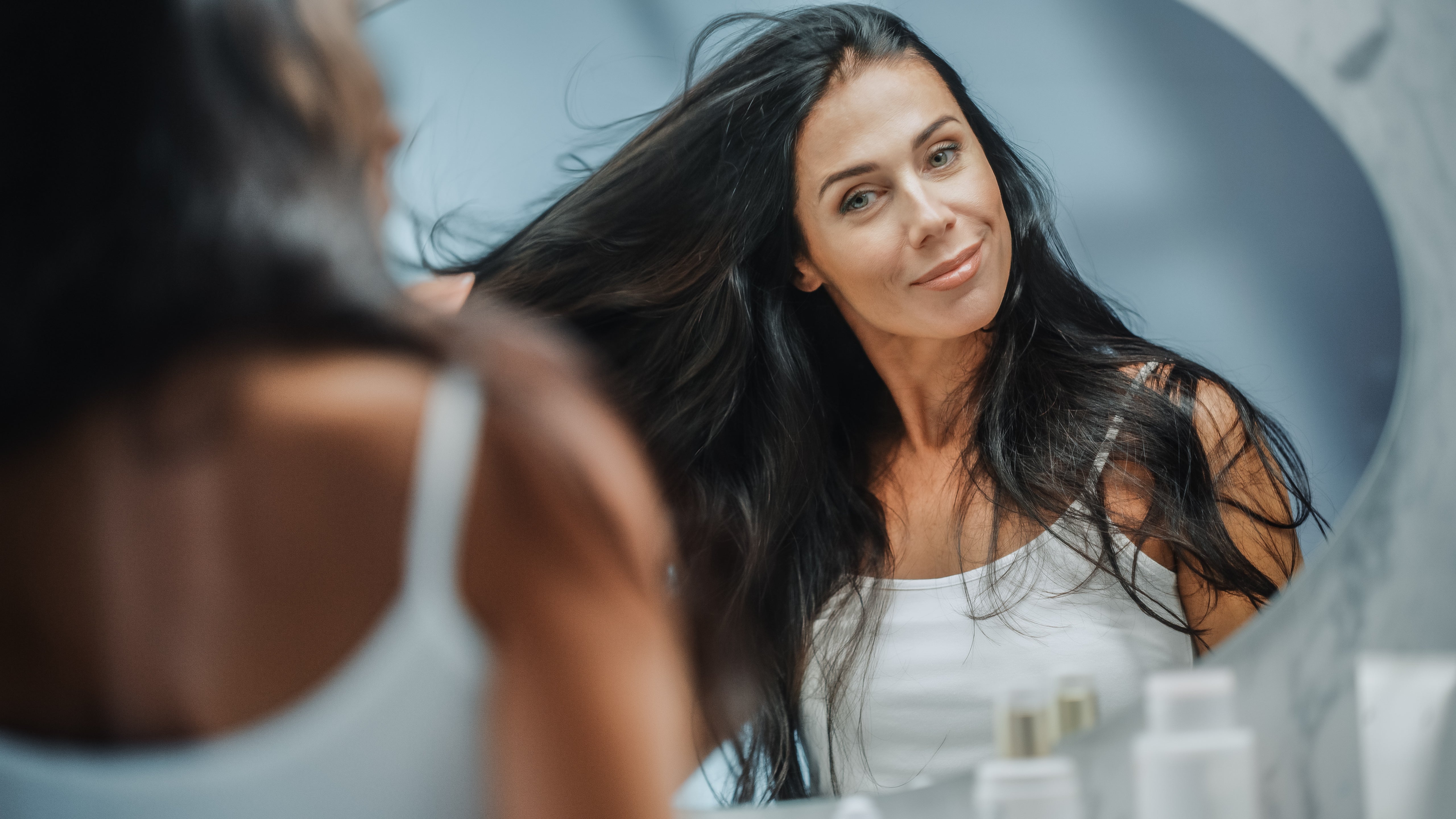 A white woman in a tank top admires her long, dark, thick and healthy hair in a mirror