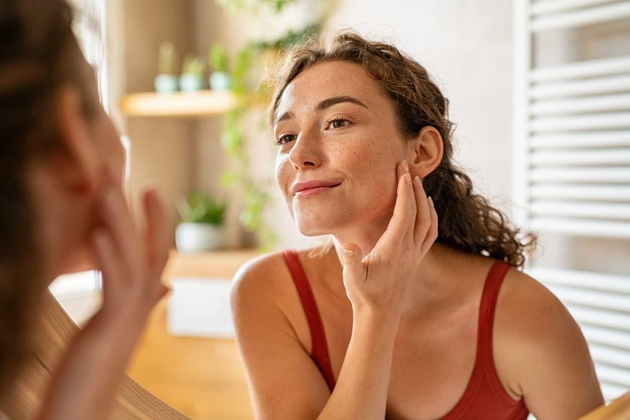 White woman smiling in a mirror as she applies skin cream to her face