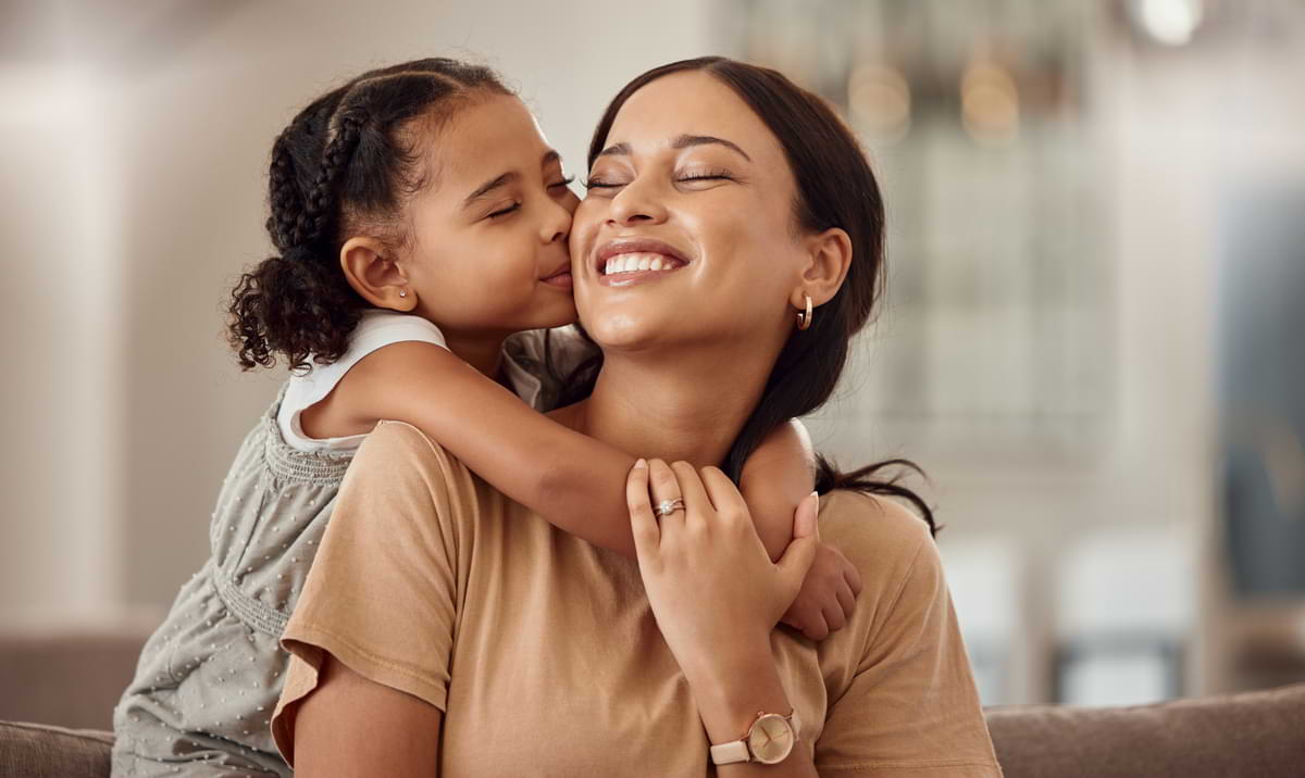 Relaxed, light-skinned mother smiles as she receives a hug and kiss from her young daughter