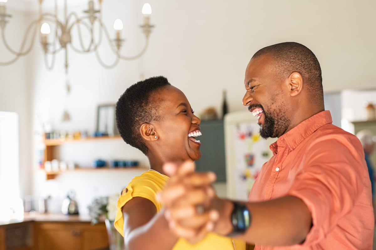 A black couple smiles at each other while dancing together in their kitchen.