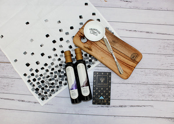 Flat lay on white washed timber with timber cheeseboard, linen tea towel, chocolate and infused olive oil bottle
