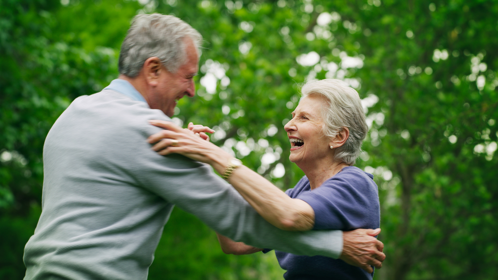 Elderly husband and wife happily dancing outdoors in front of green trees