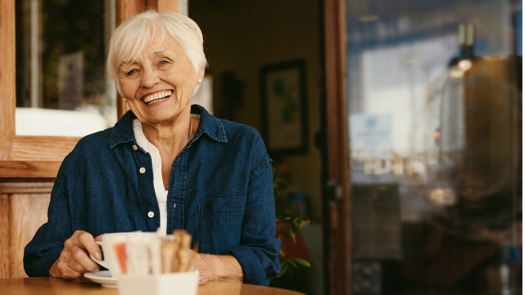 Older woman wearing a jean, buttoned-down shirt smiling and drinking tea in her kitchen