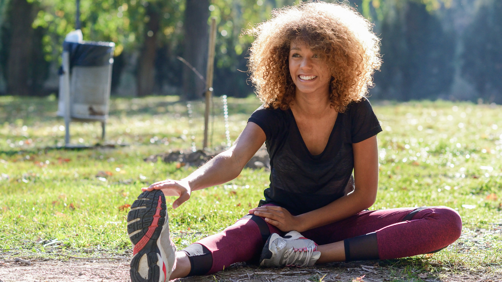 Woman in a black workout top and maroon leggings, sitting and stretching her right leg after a run