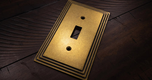 A close-up photograph of the Steps rustic brass metal wallplate laying on dark wood backdrop