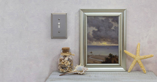 A photograph of the Georgian brushed nickel wallplate the wall of a room decorated in a Coastal decor style, and next to an ocean painting, a seashell, and a jar of shells