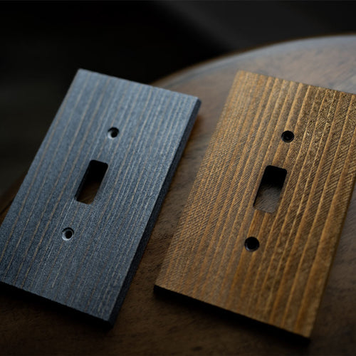 A close-up photograph of the Montana wood wallplate collection in both rustic gray and rustic brown finish options, laying on a wood table